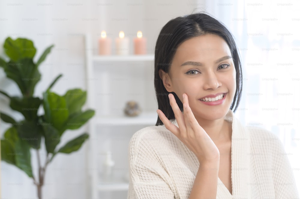 A happy beautiful woman in white bathrobe applying moisturizing cream on face , skin care and treatment concept