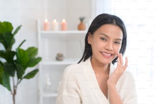 A happy beautiful woman in white bathrobe applying moisturizing cream on face , skin care and treatment concept