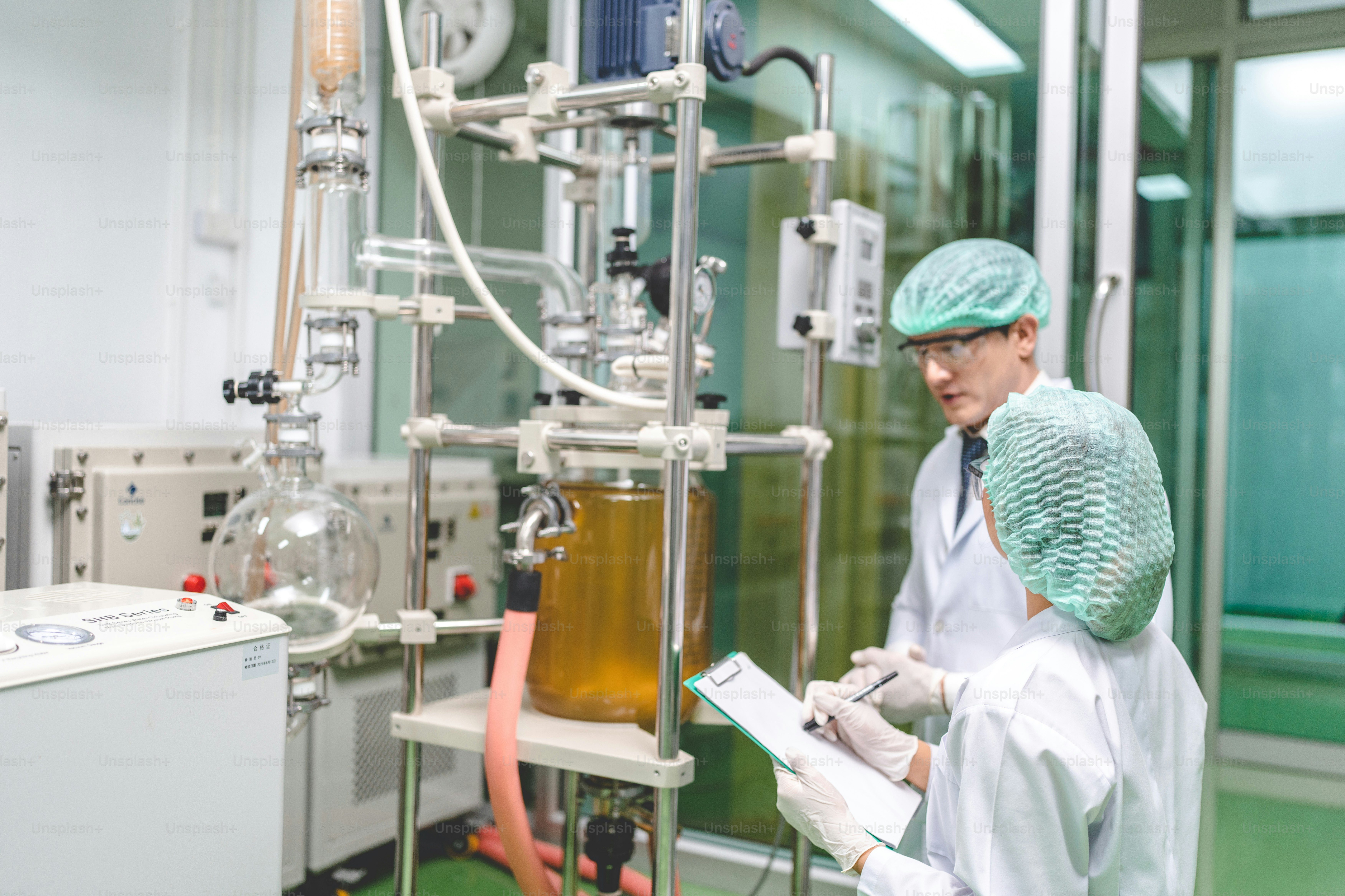 Zwei Wissenschaftler in Laborkitteln und Haarnetzen beobachten einen chemischen Reaktor in einem Labor