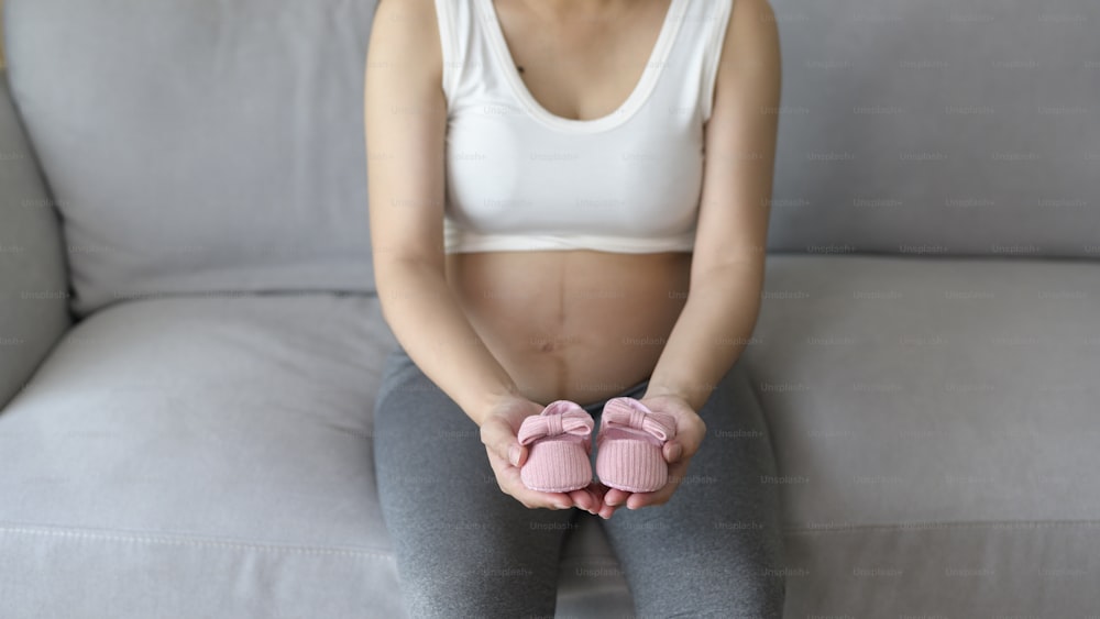 Mujer embarazada joven que pone zapatos de bebé en la barriga, familia y concepto de cuidado del embarazo