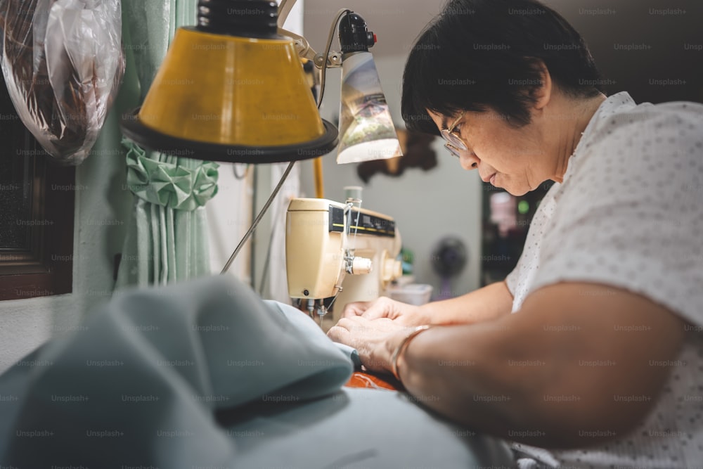 Retriement tailor southeast asian elderly crafting a cloth by sewing machine. Baby boomer generation mature working at home concept. Life moments in candid portrait authentic people.