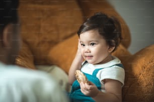 Cute little toddler girl holding circular cookie toy while sitting on floor with stuff toy in living room playing with young father at home
