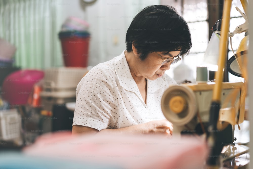 Retriement tailor southeast asian elderly crafting a cloth by sewing machine. Baby boomer generation mature working at home concept. Life moments in candid portrait authentic people.