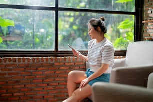 casual relaxar café estilo de vida mulher asiática sentada despreocupada emoção pacífica mão usando smartphone mídia social navegando notícias ou compras on-line com sorriso alegre ao lado da luz do sol da grande janela