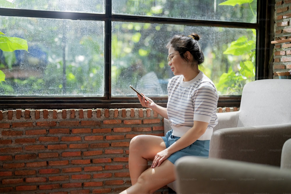 カジュアルリラックスカフェライフスタイルアジアの女性に座ってのんきで平和な感情手スマートフォンソーシャルメディアブラウジングニュースまたはオンラインショッピングを使用して、大きな窓の太陽の光の隣に陽気な笑顔で