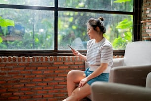 casual relax cafe lifestyle asian female woman sitting carefree peaceful emotion hand using smartphone social media browsing news or shopping online with cheerful smiling next to big window sun light