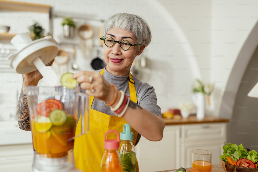 Reife lächelnde Tattoo-Frau, die Salat, Obst und Gemüse isst. Attraktive reife Frau mit frischem grünem Obstsalat zu Hause. Seniorinnenschürze steht in der Küchentheke und entspannt sich im Haus