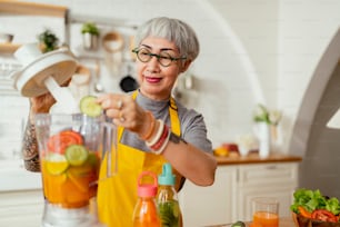 Mature smiling tattoo woman eating salad, fruits and vegetables. Attractive mature woman with fresh green fruit salad at home. Senior woman apron standing in the kitchen counter relaxing in house