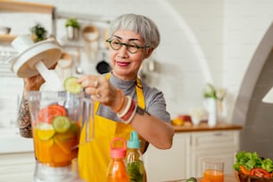 Femme tatouée souriante mature mangeant de la salade, des fruits et des légumes. Jolie femme mûre avec une salade de fruits verts frais à la maison. Tablier de femme âgée debout dans le comptoir de la cuisine se relaxant dans la maison