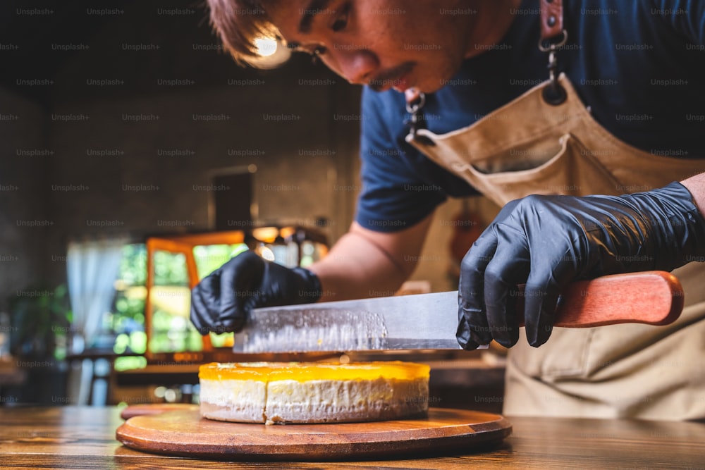 Closeup of hands of young male chef baker in unform wearing black hand gloves cutting slice of mango cheesecake using a big knife for equal sizes in pastry shop
