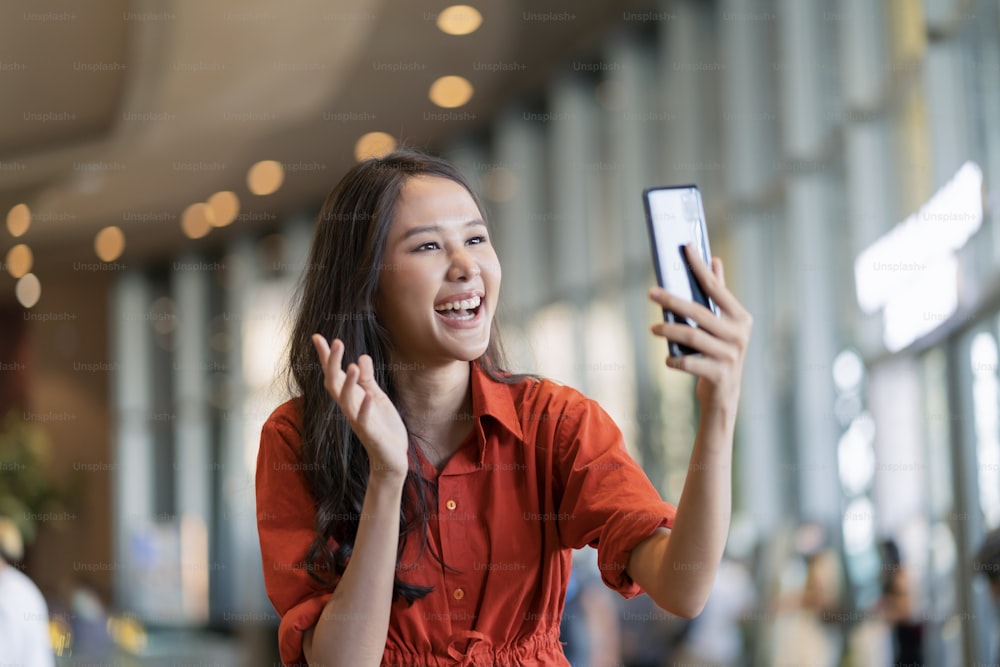 Glück Asiatische Frau Lächeln Hand Welle Geben Sie einen Kuss an die Familie per Videoanruf Smartphone-Gerät Social Distancing Begrüßung mit verschwommenem Mall-Hintergrund Neuer normaler Lebensstil