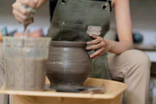 Nahaufnahme des asiatischen Künstlers Keramiker weibliche Hände, die an der Töpferscheibe arbeiten, asiatische weibliche Skulptur Frau mit nassen schmutzigen Händen, die eine Tonvase auf der Töpferscheibe im Werkstattstudio formen