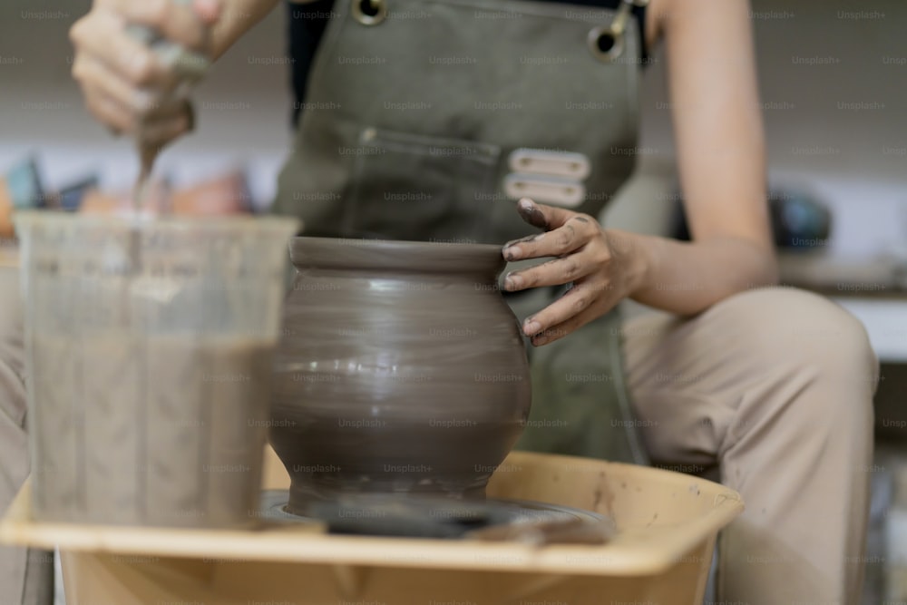 Primer plano de las manos femeninas ceramistas del artista asiático que trabajan en la rueda de alfarero, mujer asiática de la escultura femenina con las manos sucias mojadas que dan forma al jarrón de arcilla en la rueda de alfarería en el estudio del taller