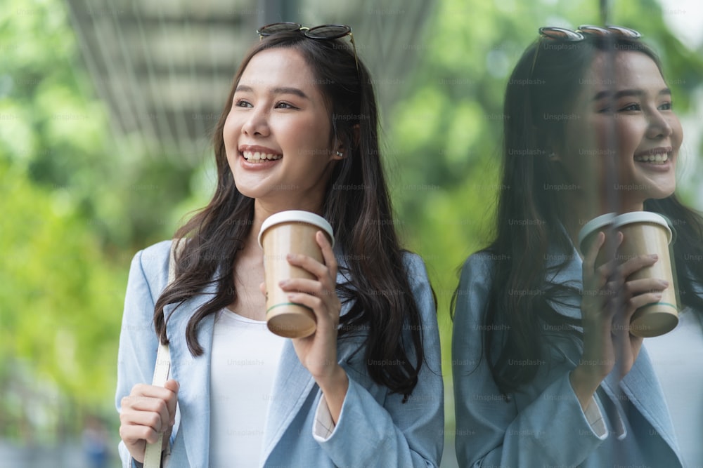 Asiática femenina casual relajarse nómada digital freelance expatriado programador inteligente casual tela caminando en la acera ciudad urbana con sosteniendo taza de café sonriendo alegre sentimiento positivo downshifting estilo de vida
