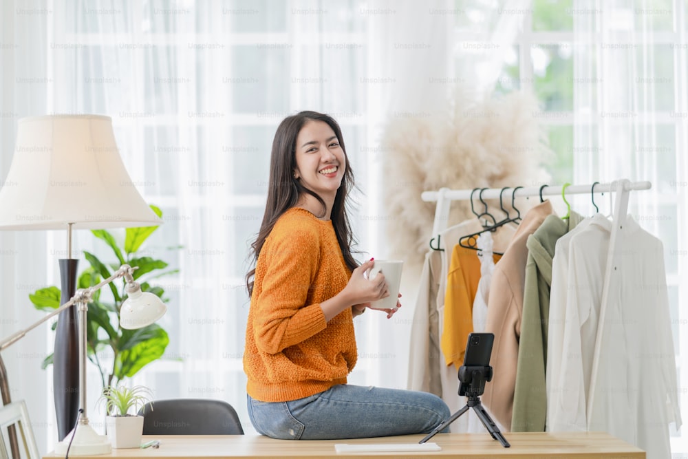 intelligente attraente sorridente asiatico imprenditrice imprenditore mano tenere tazza di caffè ritratto girato guardando la macchina fotografica con fiducioso, donna seduta sul tavolo a casa studio