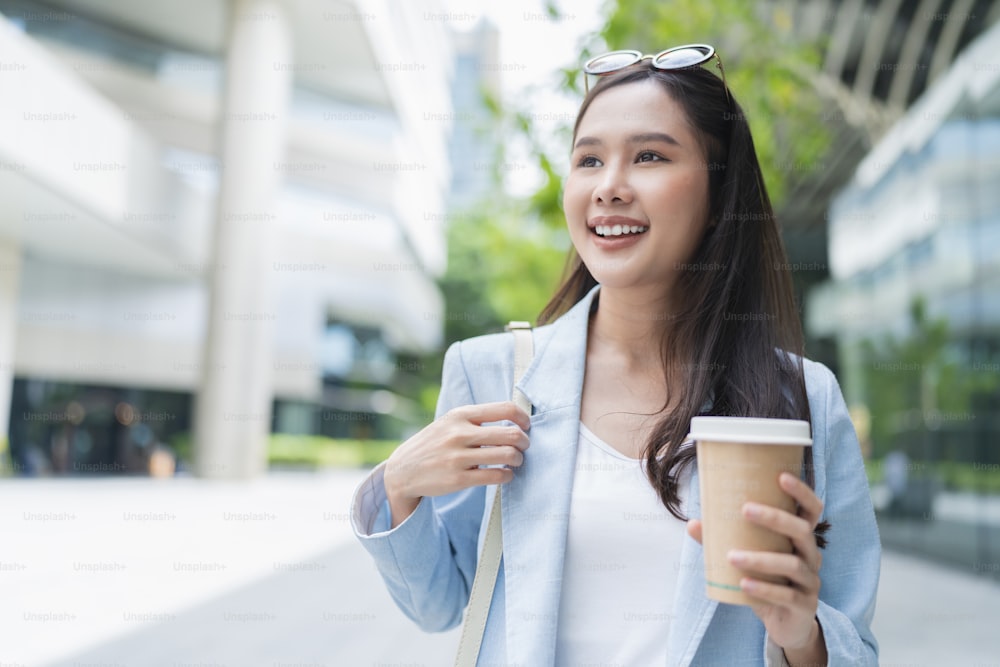 Mujer asiática relajada casual nómada digital freelance expatriada programadora inteligente ropa casual caminando en la acera de la ciudad urbana con una taza de café sonriendo alegre sentimiento positivo cambio de estilo de vida