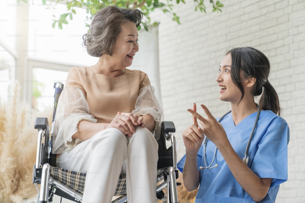 Asiatische junge Pflegekraft kümmert sich um ihre ältere Patientin in der Seniorentagesstätte Behinderte Patientin im Rollstuhl im Krankenhaus im Gespräch mit einer freundlichen Krankenschwester und sieht fröhlich aus Krankenschwester Ältere Patientin