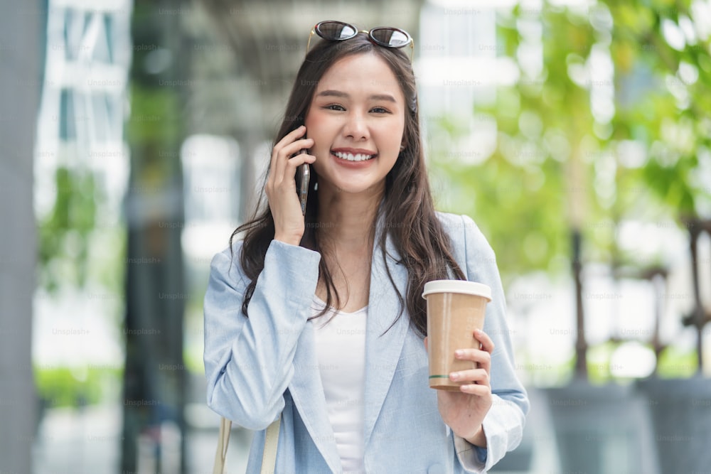 Asian female casual relax digital nomad freelance expat programer smart casual cloth walking on sidewalk urban city with holding smartphone smiling cheerful positive feeling downshifting lifestyle
