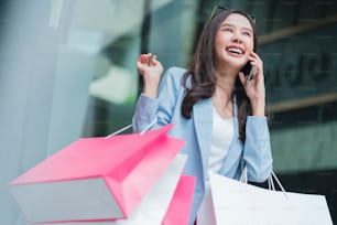 attractive asian woman pleased positive casual shopping hand hold paper bags and smartphone enjoy purchase cloths shopping buy sales discount at center mall store shopfront corridor department store