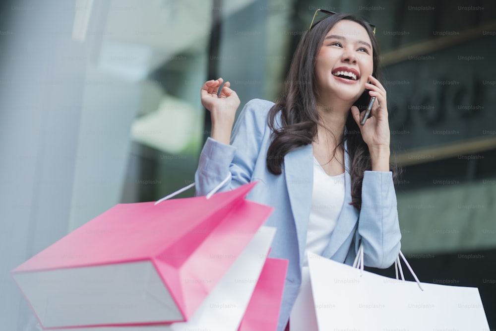 attractive asian woman pleased positive casual shopping hand hold paper bags and smartphone enjoy purchase cloths shopping buy sales discount at center mall store shopfront corridor department store