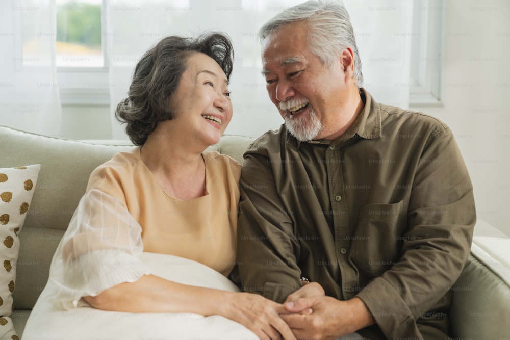 古いシニアアジアの退職年齢は、自宅で一緒にカップルのウェルネスライフスタイルと結婚し、老人はリビングルームの家のインテリアの背景のソファで愛と絆と一緒に笑顔を笑います