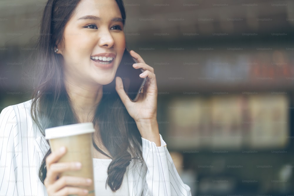 attractive asian female adult business woman formal cloth happiness smiling conversation discuss talk hand hold smartphone and coffee cup communication with reflect view through cafe window shopfront