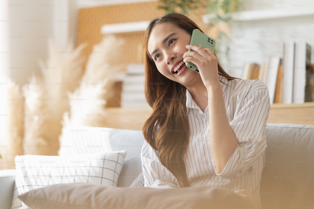 felicidad alegre sonriente joven adulta mujer asiática usa ropa casual conversación de mano en el teléfono inteligente en el sofá en casa cuarentena remota período de bloqueo fondo interior de la casa