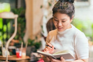 캐주얼 휴식 아시아 여성 프리랜서 디지털 유목민 손 쓰기 스케치 책 아이디어 카페에서 그녀의 새로운 프로젝트로 주목 커피 숍 창 유리를 통해 촬영, 아시아 여성 카페에서 작업 쾌활한 느낌