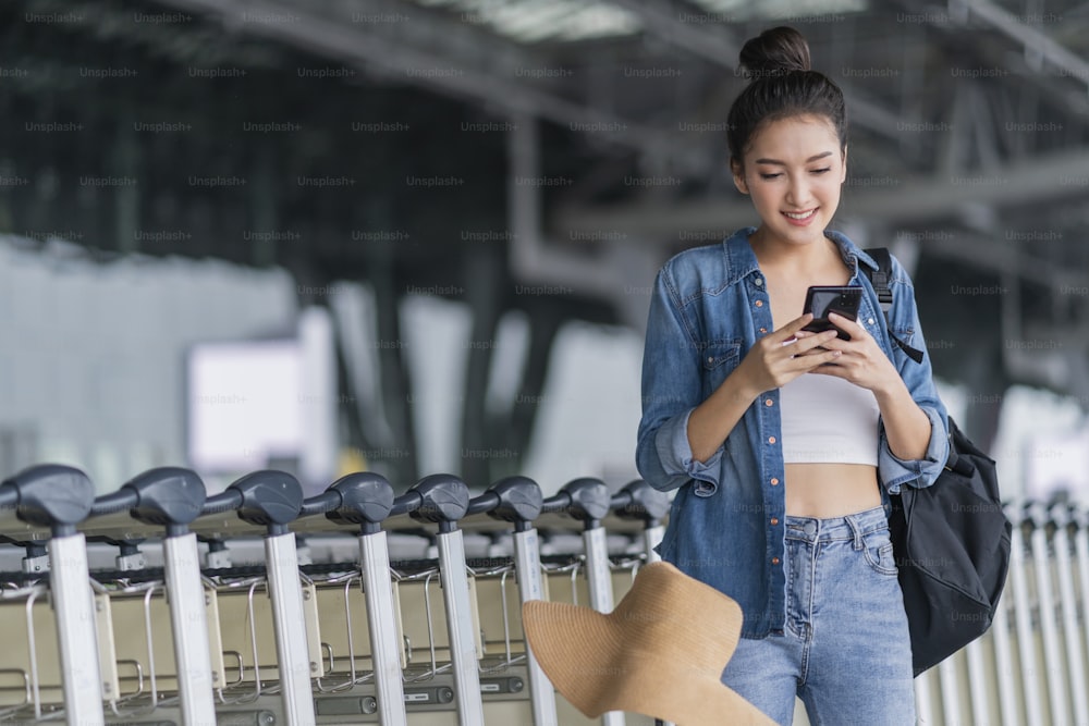 Expat Digital Nomad Reisen und Arbeit, asiatische fröhliche weibliche lässige Kleidung Gehen während der Verwendung von Smartphone-Buchung Check-in Flug Online-Ticket-Informationsanwendung am Flughafen Terminal Reisekonzept