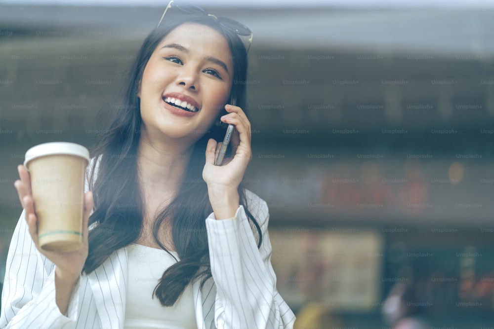 attraente asiatico femmina adulto donna d'affari formale panno felicità sorridente conversazione discutere parlare tenere la mano smartphone e tazza di caffè comunicazione con riflettere vista attraverso la finestra del caffè vetrina