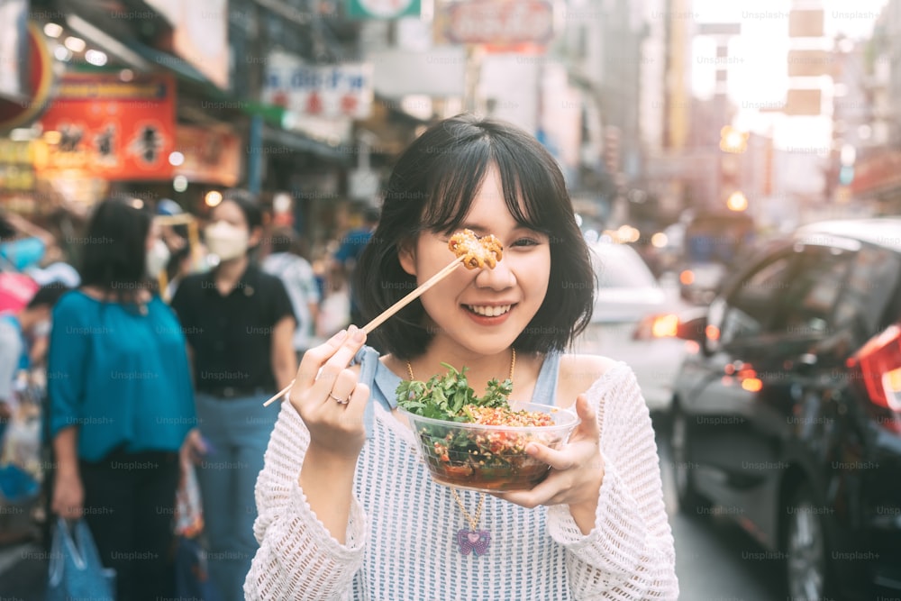 Poeple旅行と屋台の食べ物のコンセプトを食べる。東南アジアのチャイナタウン市場でスパイシーなイカのグリルを保持している幸せな若い大人のアジアの食通の女性。