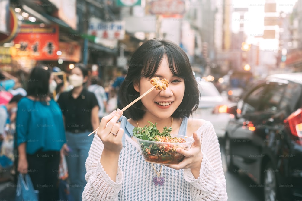 Poeple viajar e comer conceito de comida de rua. Feliz jovem adulto asiático foodie mulher segurando lula grelhada picante no mercado de Chinatown do sudeste da Ásia.