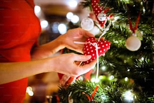 Unrecognizable young woman in red dress in front of illuminated Christmas tree inside in her house decorating it.