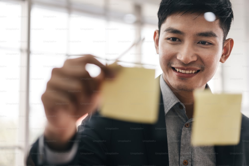 Happy smile positive adult business asian man. Write a creative idea project in post it and stick on a glass board. Professional persona manager working at workplace. Modern office window with light.