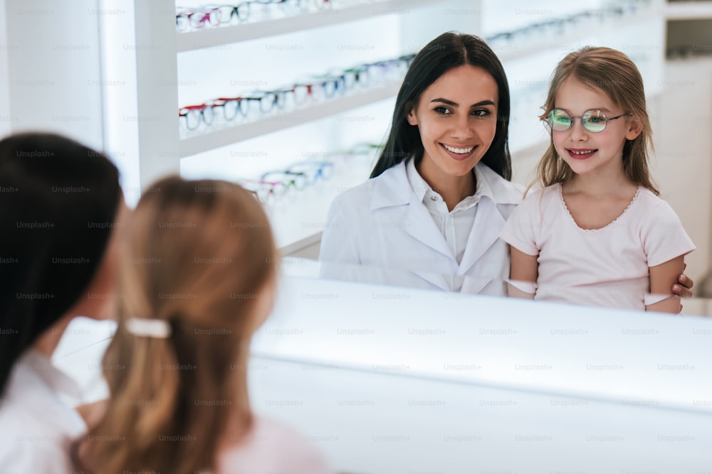 Attractive female doctor ophthalmologist is helping little cute girl to choose the best eyeglasses in modern ophthalmology clinic.
