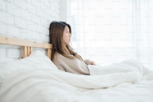 Happy beautiful young Asian woman waking up in morning, sitting on bed, stretching in cozy bedroom, looking through window. Funny asian woman after wake up. Asia woman is stretching and smiling.