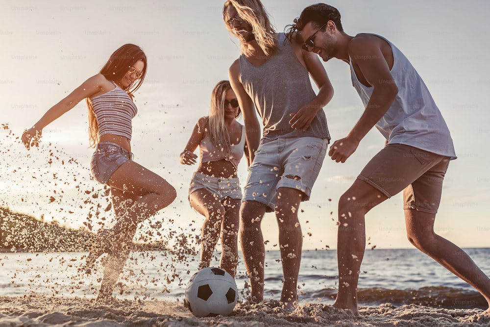 Gruppo di giovani amici attraenti si divertono sulla spiaggia e giocano a calcio