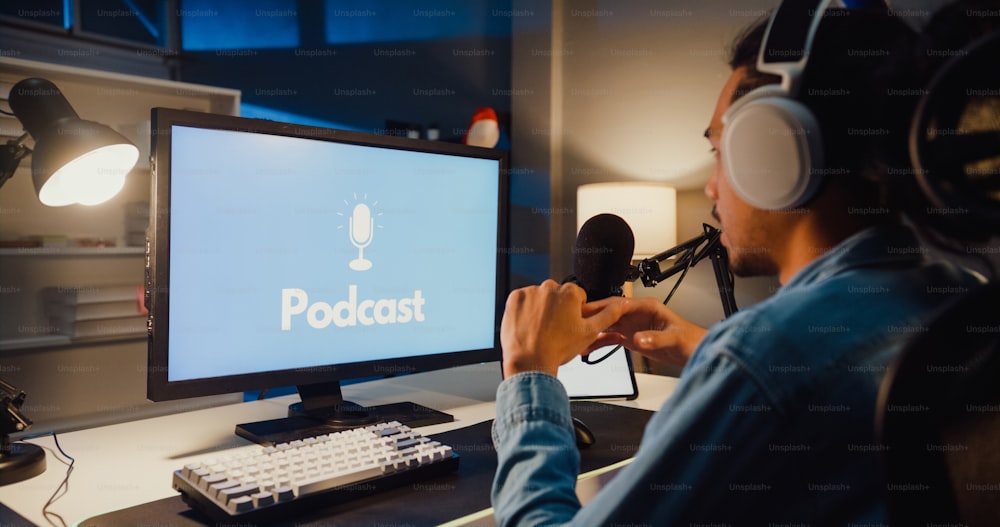 Happy Asia Guy Blogger Musik-Influencer nehmen einen Podcast auf dem Computer mit Kopfhörern und Mikrofon sprechen mit dem Publikum im Wohnzimmer Heimstudio in der Nacht. Bleiben Sie zu Hause, Content-Ersteller-Konzept.