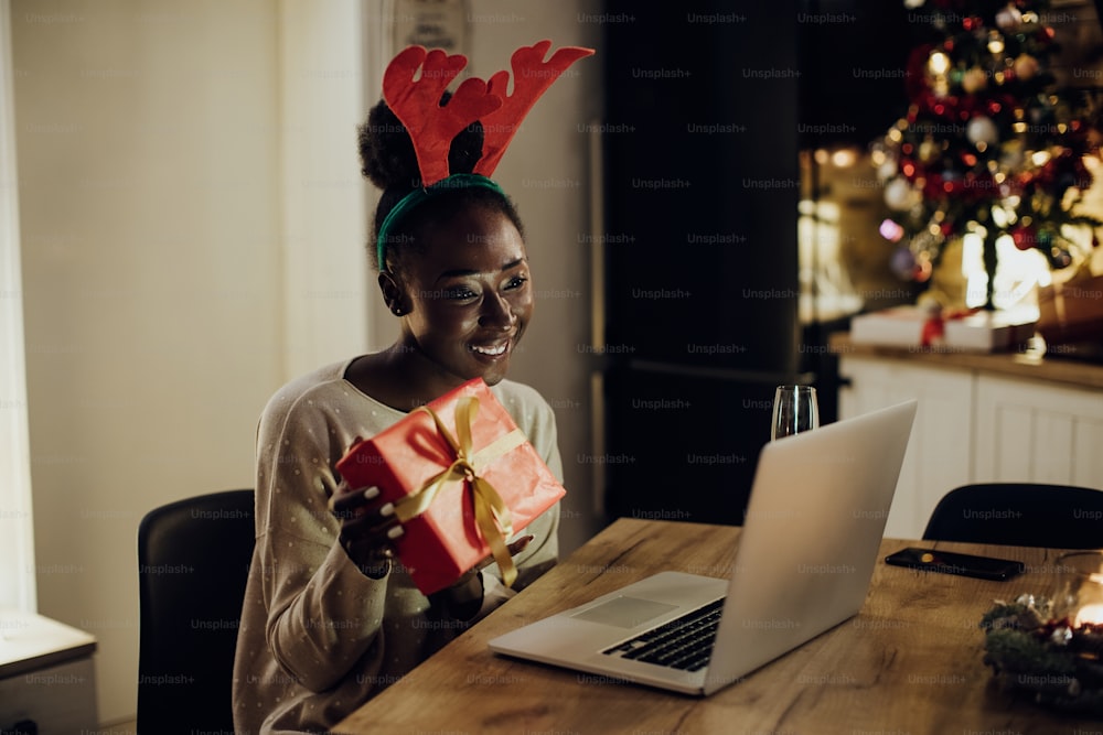 Donna afroamericana felice che mostra un regalo mentre effettua una videochiamata tramite laptop la sera di Natale a casa.