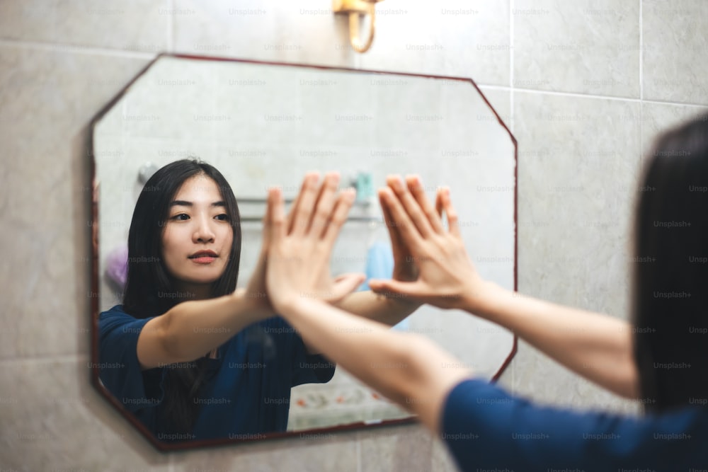 Young adult smile asian woman practice self talk conversation with mirror. Mental health in bathroom at home. Healthy lifestyle after wake up life with satisfaction concept.