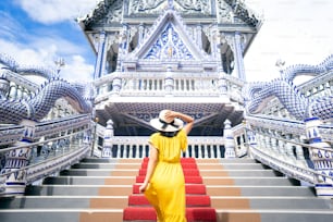 Back view of solo asian woman wear yellow dress and hat. Walking to blue temple on day. Outdoor culture local asia travel. Wanderlust destination in Thailand tourism concept.