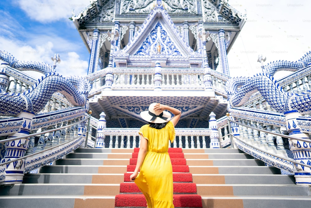 Back view of solo asian woman wear yellow dress and hat. Walking to blue temple on day. Outdoor culture local asia travel. Wanderlust destination in Thailand tourism concept.