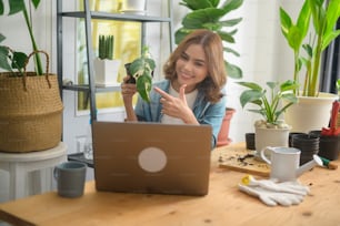 A young woman entrepreneur working with laptop presents houseplants during online live stream at home, selling online concept
