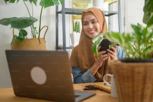 Una joven empresaria musulmana que trabaja con una computadora portátil presenta plantas de interior durante la transmisión en vivo en línea en casa, vendiendo el concepto en línea