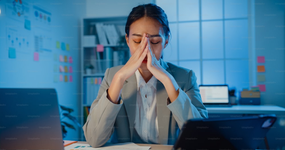 Jeune femme d’affaires asiatique assise avec un ordinateur portable et une tablette sur le bureau, se frottant les yeux, ressent la douleur et la fatigue du surmenage au bureau la nuit. Les femmes souffrent du syndrome du bureau, du travail de longue durée, de la santé mentale sur le lieu de travail.