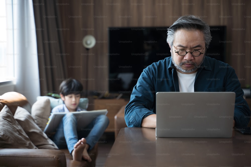 息子と一緒に自宅でノートパソコンを使って仕事をしている中年アジア人男性が、背中に絵を描いています。