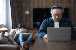 Hombre asiático de mediana edad que usa una computadora portátil para trabajar desde casa con su hijo hace un dibujo en la parte posterior.