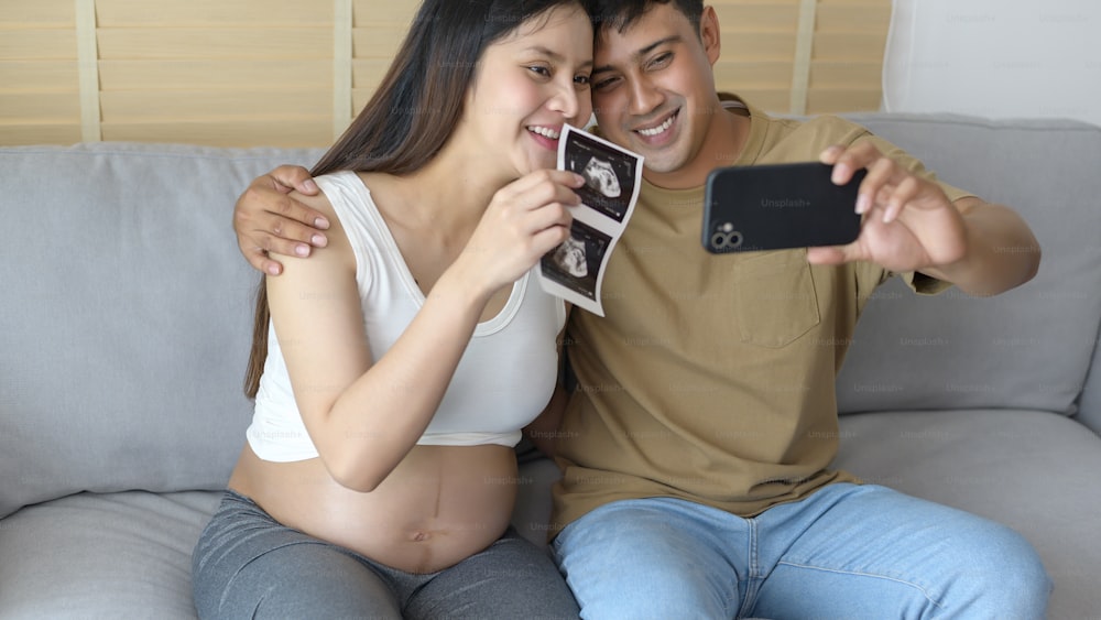 Young pregnant woman with husband embracing and video call with family and friends by smartphone on social media, family and pregnancy care concept