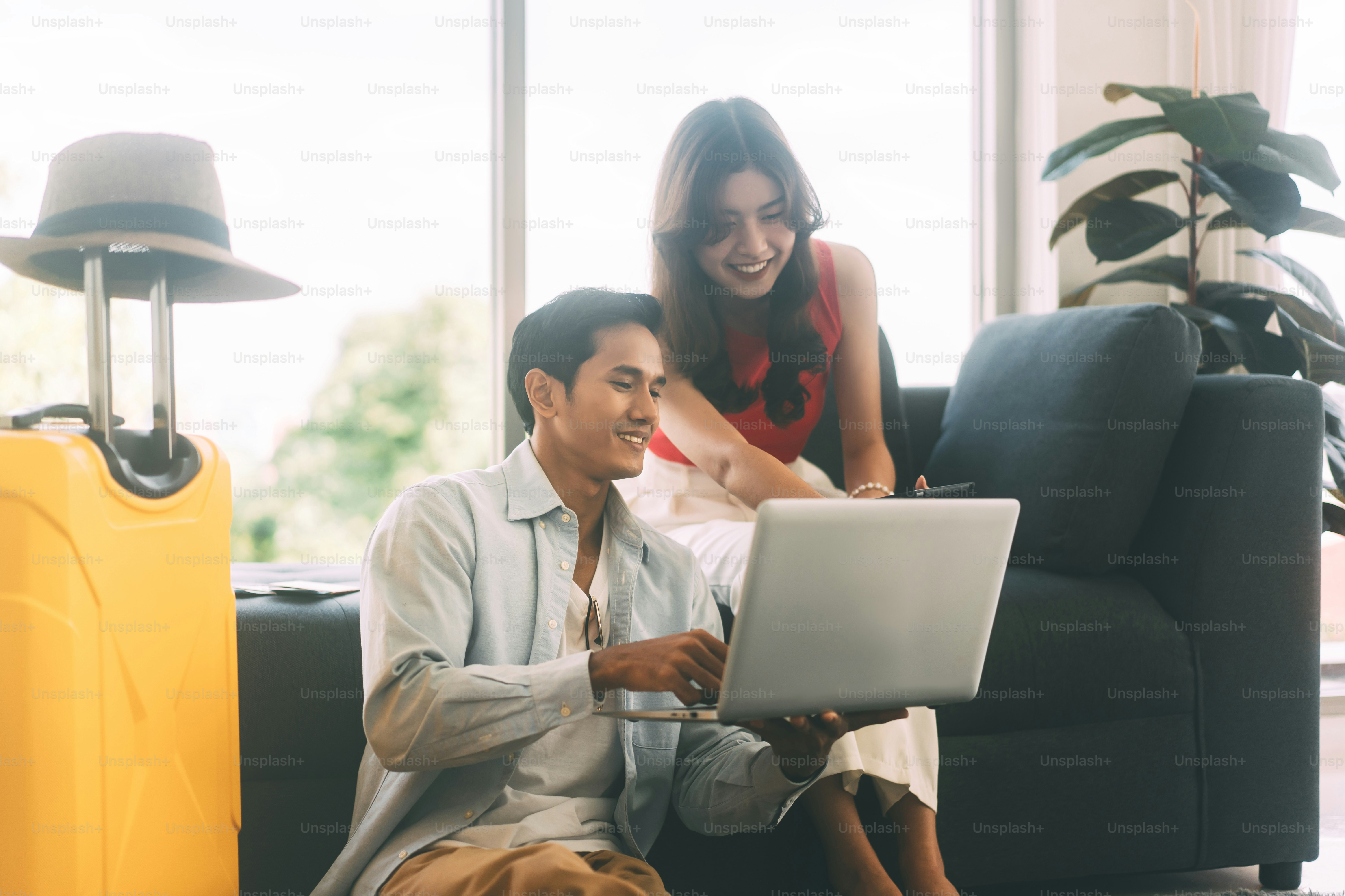 Young adult southeast asian couple using laptop for getting ready for holidays travel trip. Living at home sitting on sofa with hat and luggage. Window day light background.