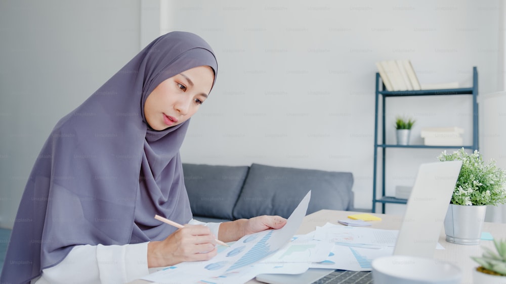Schöne asiatische muslimische Dame in Kopftuch-Freizeitkleidung mit Laptop im Wohnzimmer im Haus. Fernarbeit von zu Hause aus, neuer normaler Lebensstil, soziale Distanzierung, Quarantäne zur Prävention von Coronaviren.